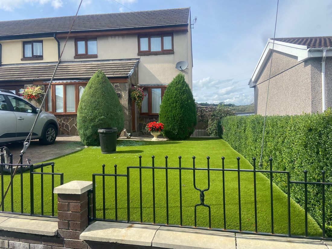 Neat green artificial lawn in a small front garden with black railings, two cone-shaped conifers and a hedge