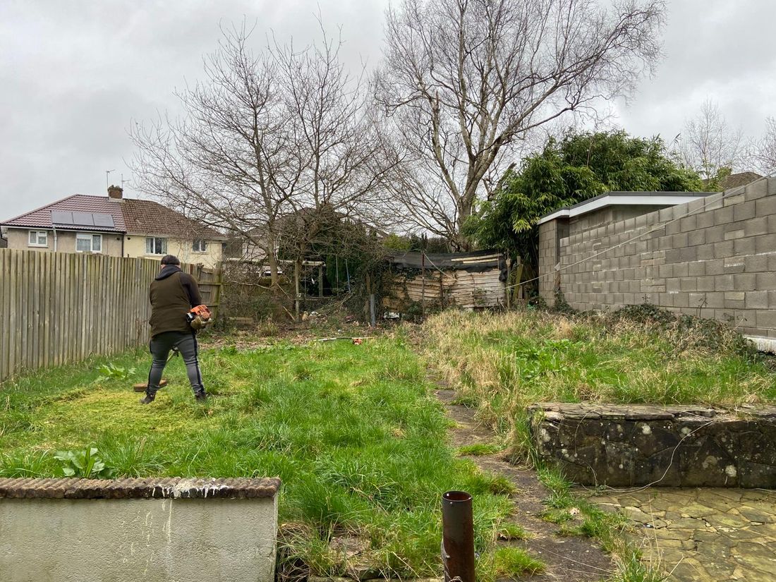 Person using a brushcutter on a long-overgrown back garden with a block wall, timber fence and bare trees