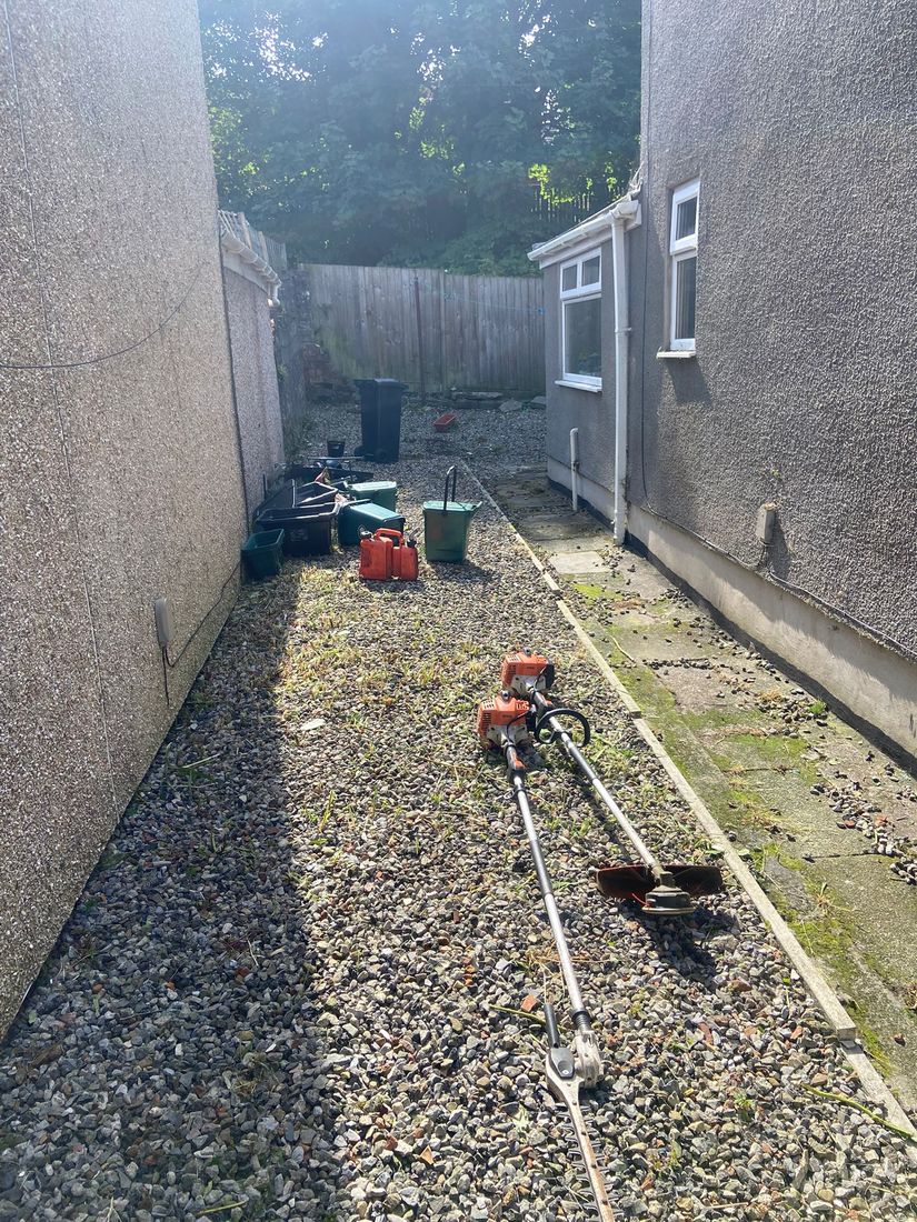 Gravel side passage between rendered house walls with petrol trimmers, fuel cans, buckets and a wheelie bin