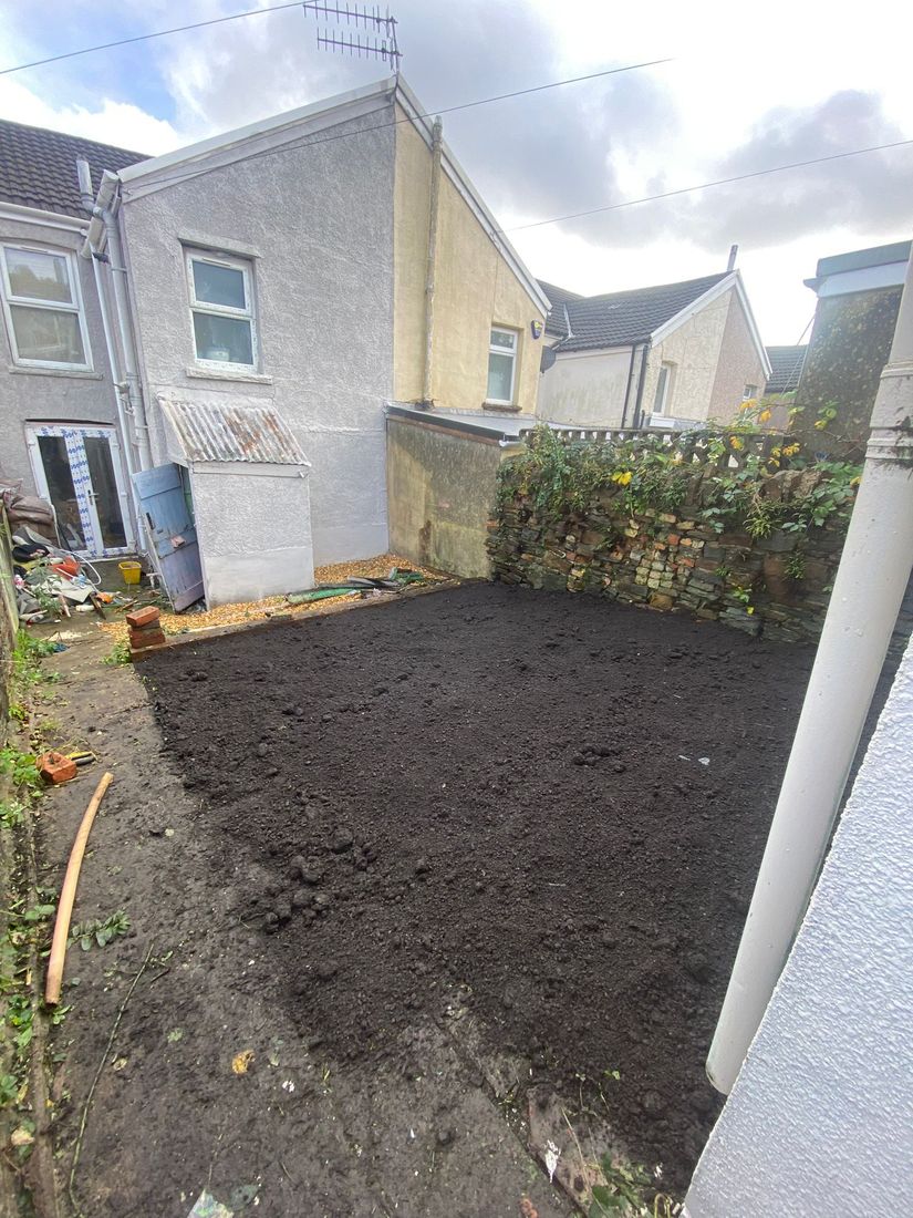 Small back garden after clearing, with flat dark soil beside a stone wall and the back of a terraced house