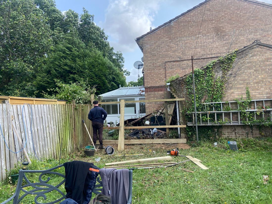 Person standing by newly set timber fence posts and rails beside an older fence and an ivy-covered brick house