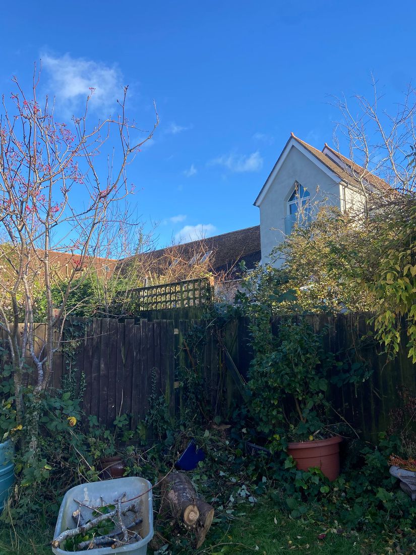 Fence line with a cut log, a barrow of branches and a house behind
