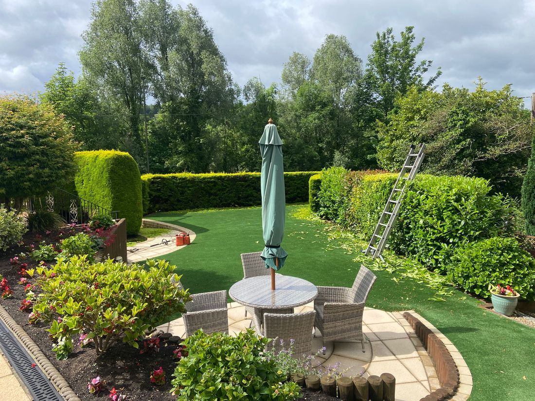 Neatly clipped hedges around a green lawn and round patio with a table, chairs and a ladder