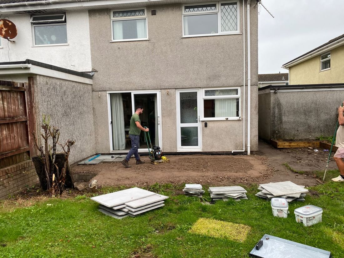 Man running a plate compactor over bare levelled soil with stacks of new paving slabs on the lawn
