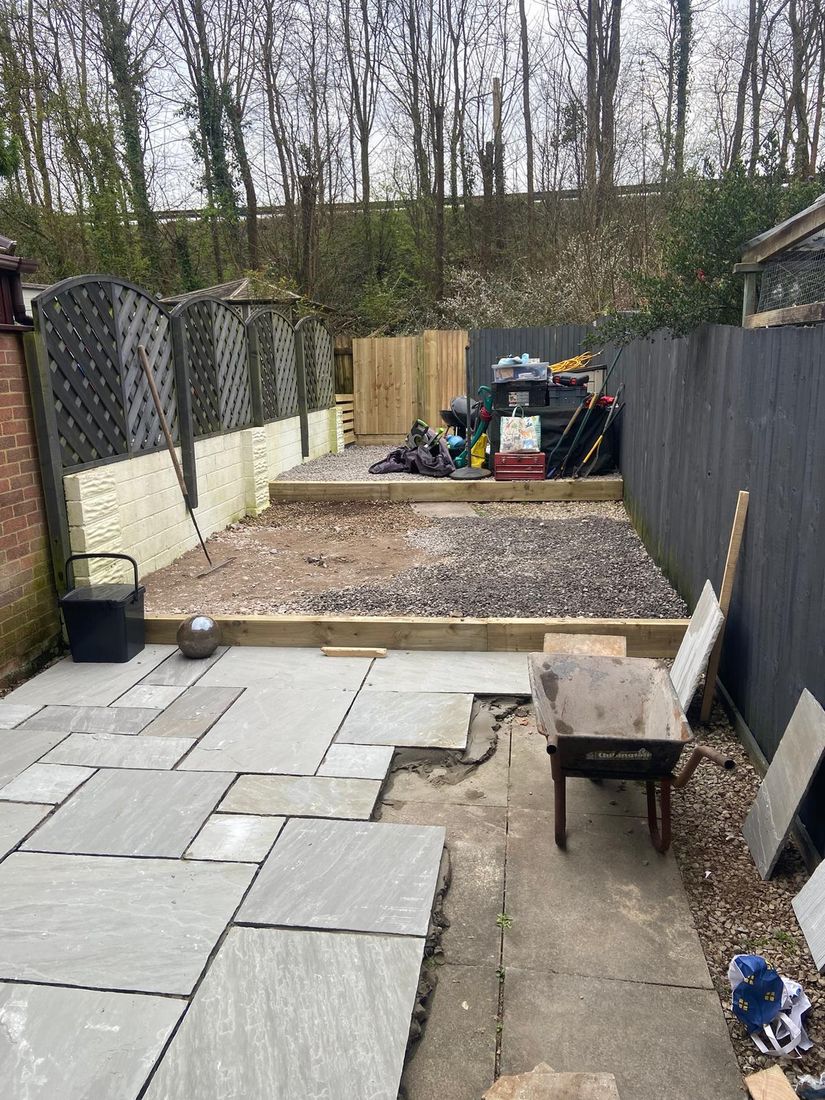 Grey paving slabs laid in a narrow garden with a wheelbarrow, timber edging and gravel beyond